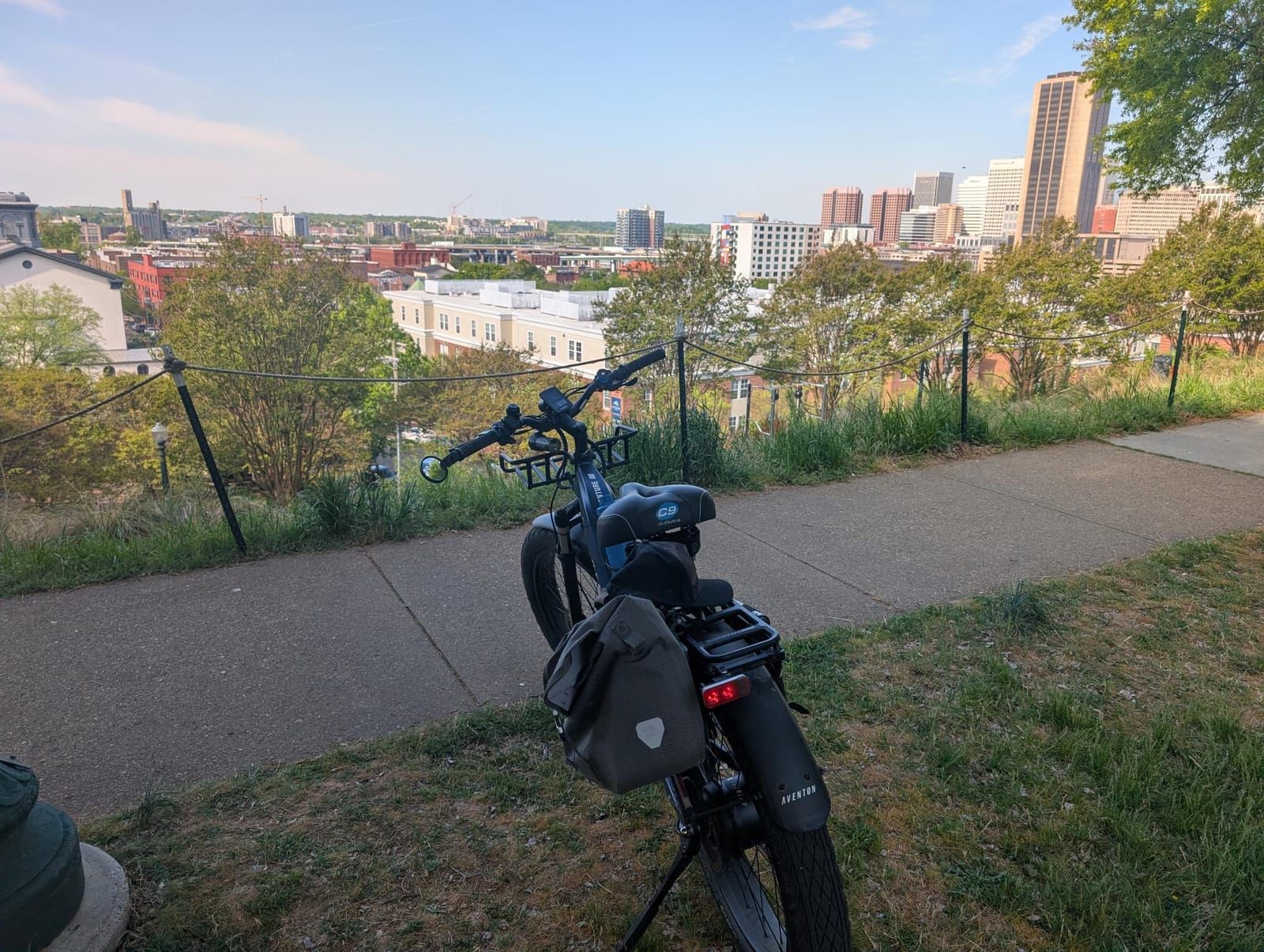 An electric bike parked on a hillside overlook with a sprawling cityscape in the background, featuring downtown buildings, residential areas, and green vegetation along the observation point.