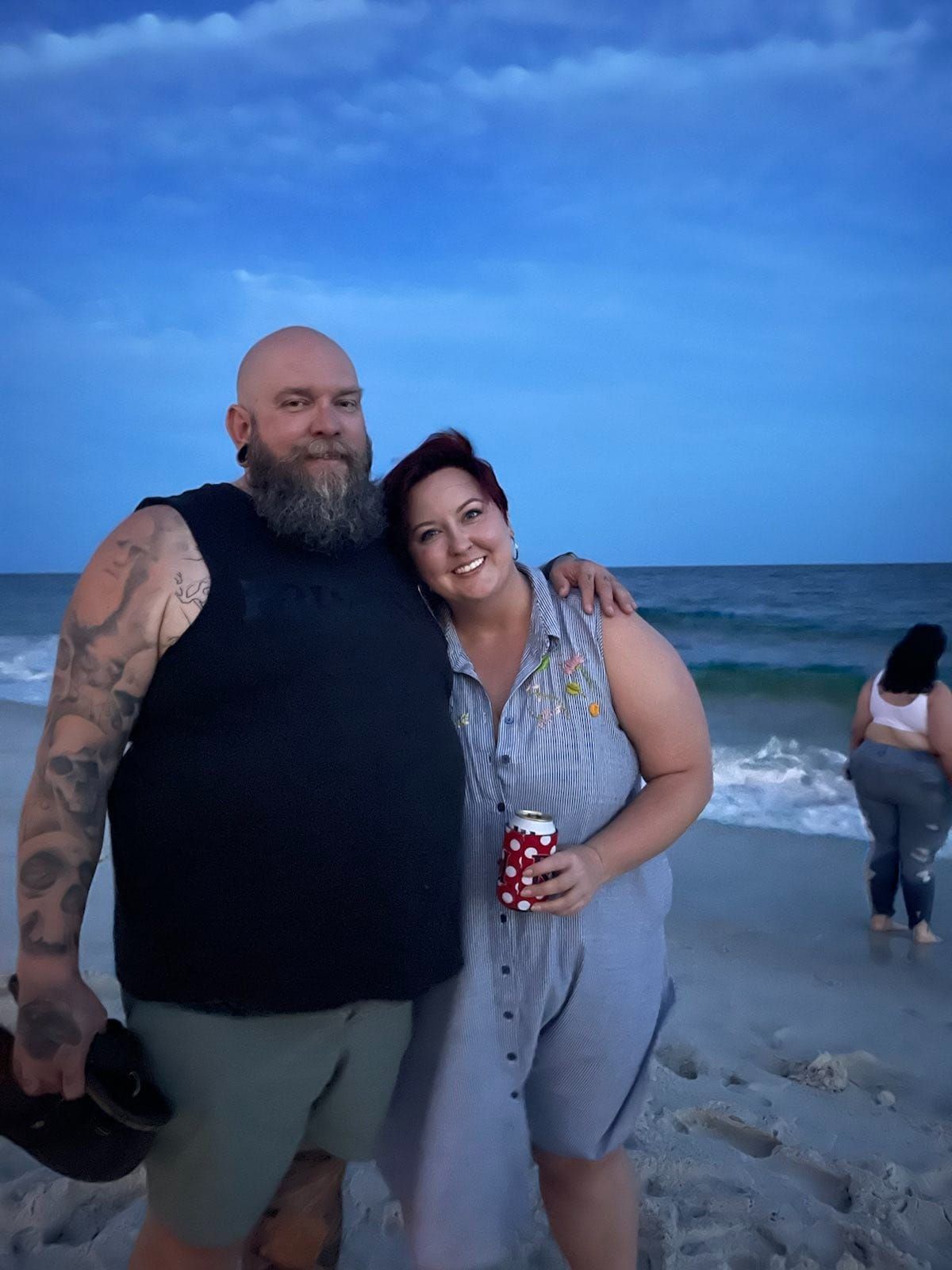 Photo of Mike and Rachelle on the evening beach. Mike wears a black cut off T and green shorts. Rachelle is holding a drink and wears a blue long dress.