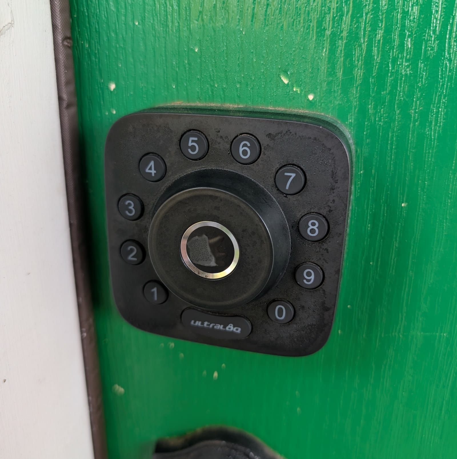 A black rotary dial keypad mounted on a bright green wooden surface. The keypad features numbered buttons 0-9 arranged in a circular pattern around a central rotating dial, with 'UltraLoo' branding visible at the bottom.
