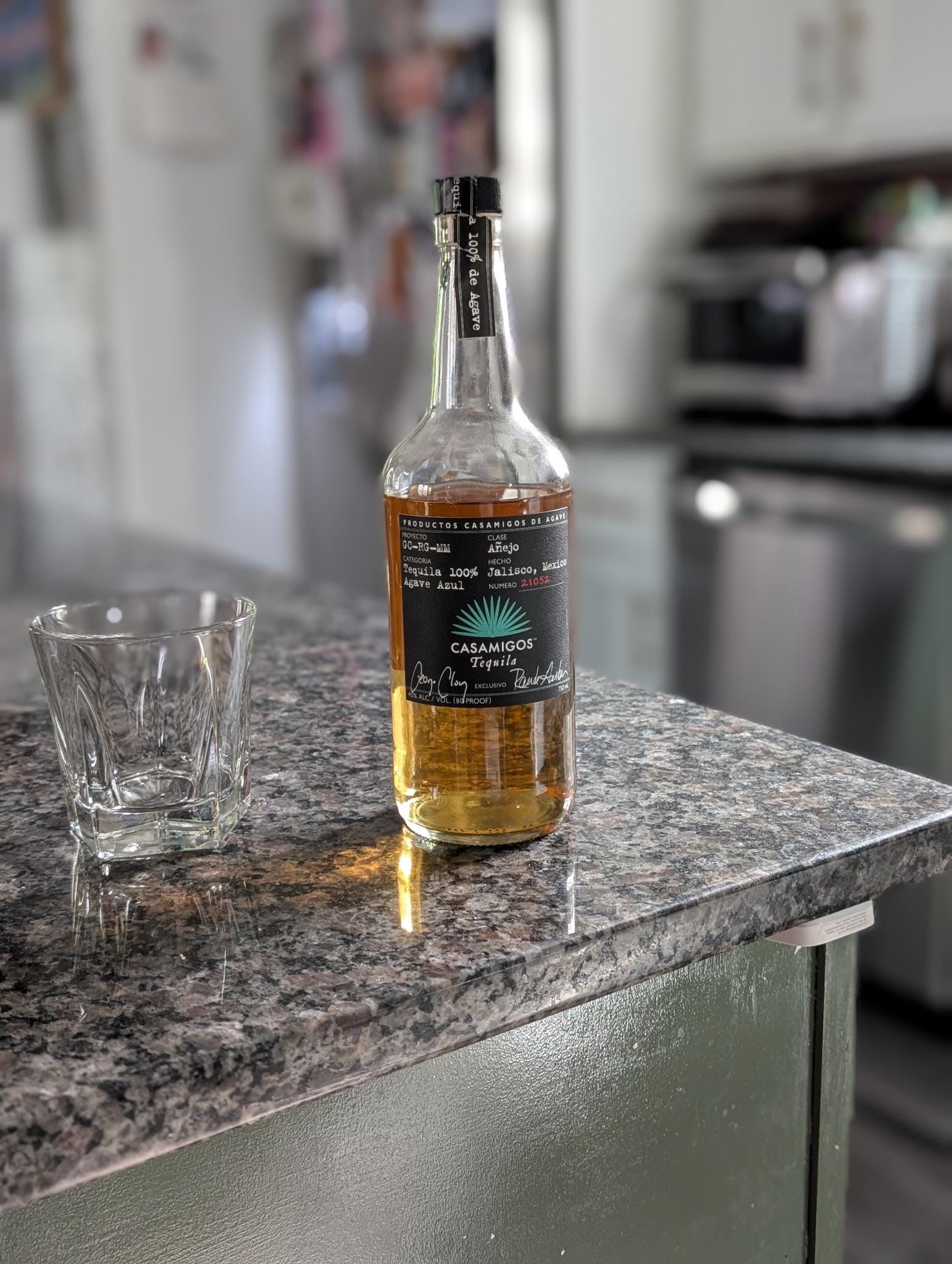 A bottle of Casamigos Tequila with a black label and gold liquid stands on a dark granite countertop next to an empty glass in a modern kitchen setting.
