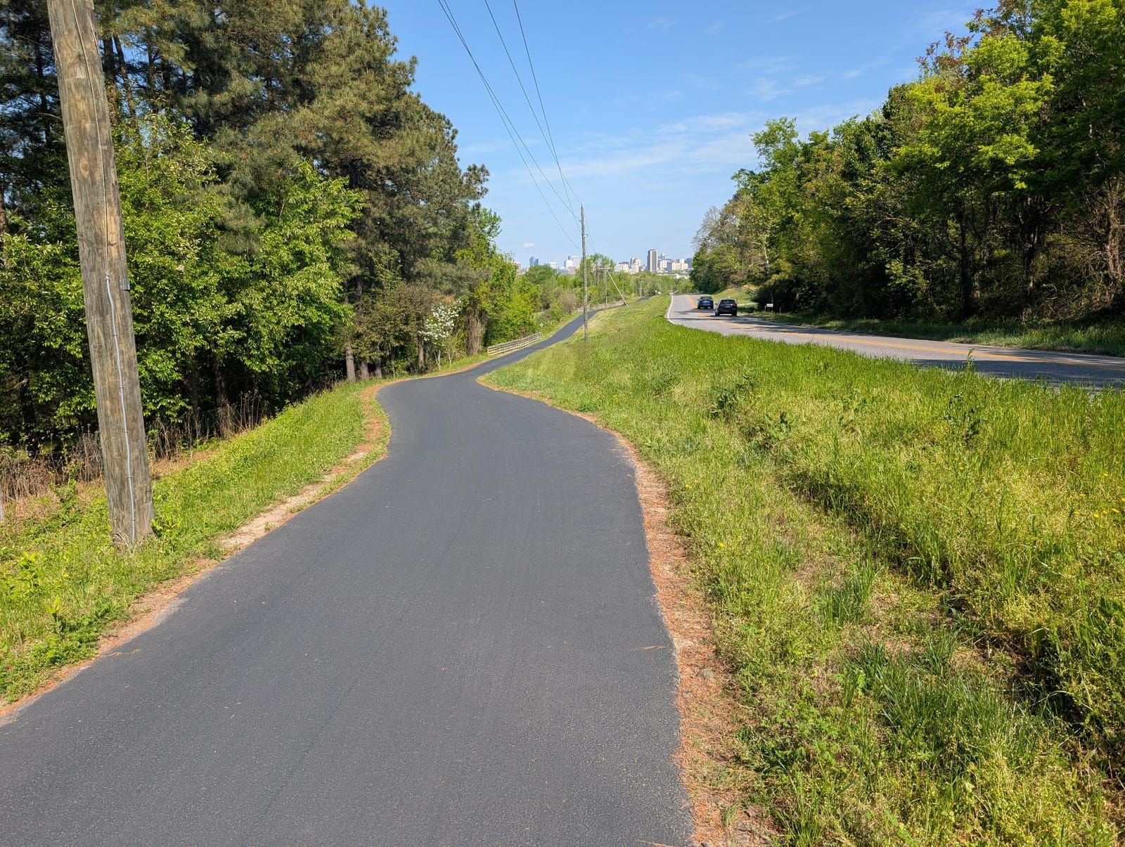 A winding asphalt path curves through a grassy, tree-lined area with a city skyline visible in the distance and a road with vehicles parallel to the path on the right.