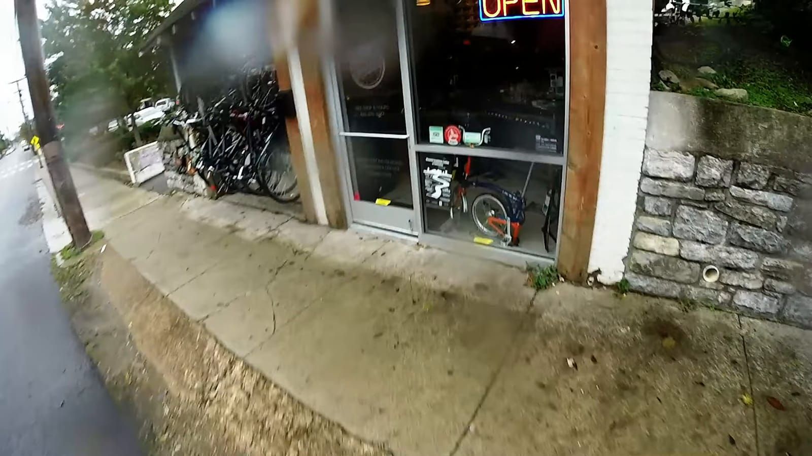 Helmet-cam view riding past a bike shop storefront with an OPEN neon sign, bicycles parked outside and displayed in the window, along a cracked sidewalk beside a stone wall.