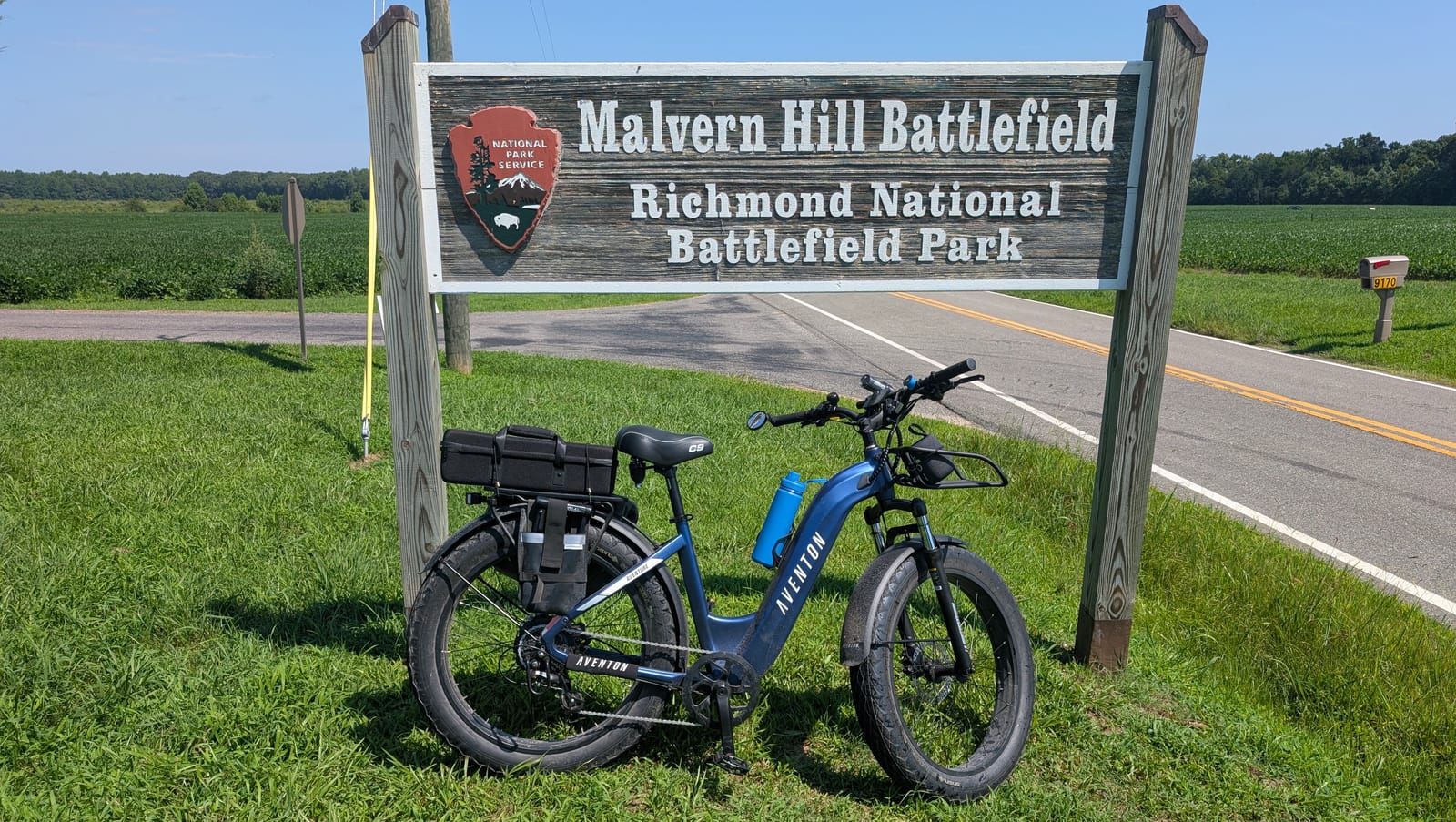 A blue Aventon electric bike with cargo bags stands on grass in front of the Malvern Hill Battlefield sign for Richmond National Battlefield Park, with a rural road and green fields visible in the background.