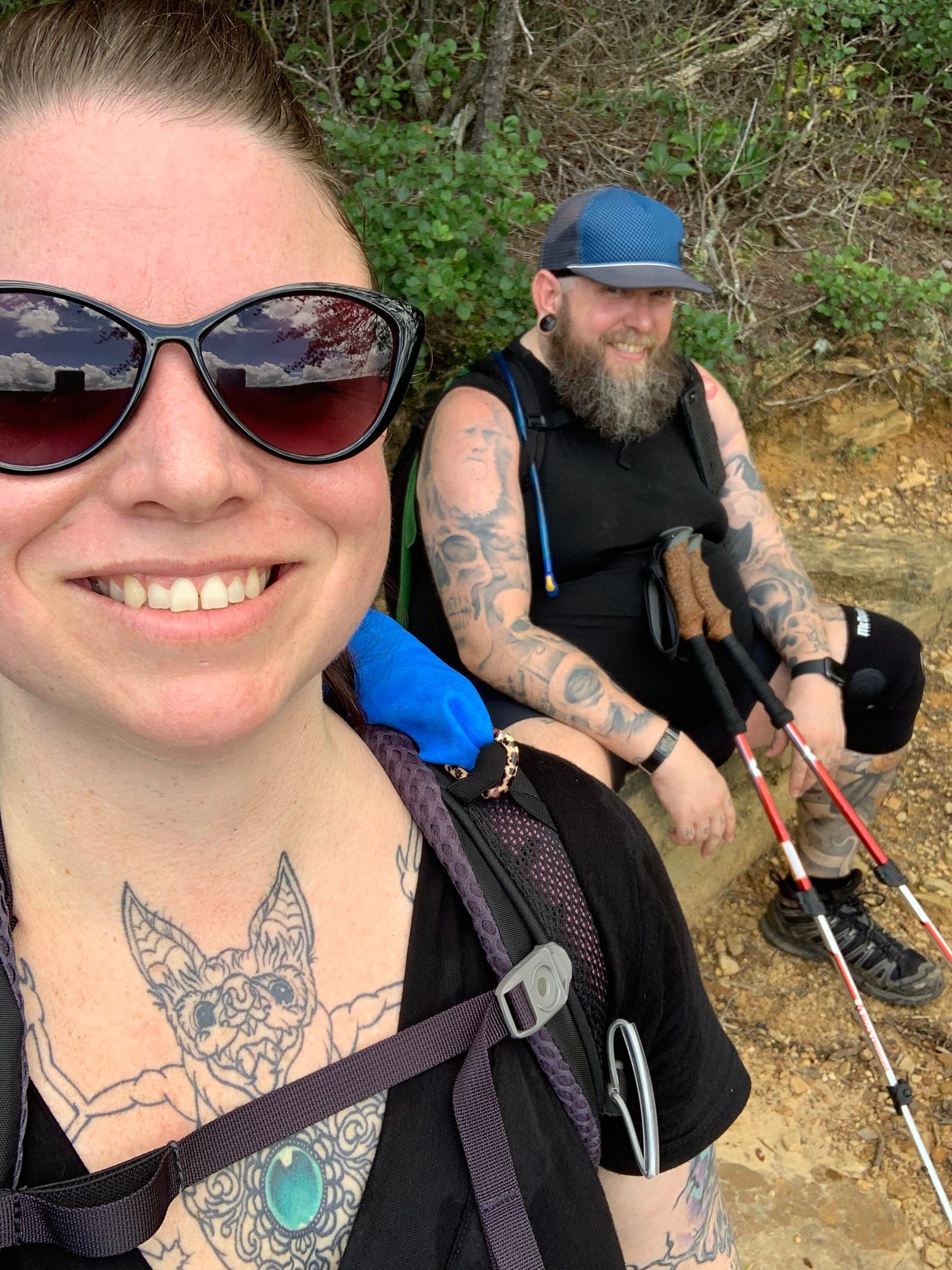 Meredith taking a smiling selfie with Mike in the background take a break on a rock and smiling at the camera.