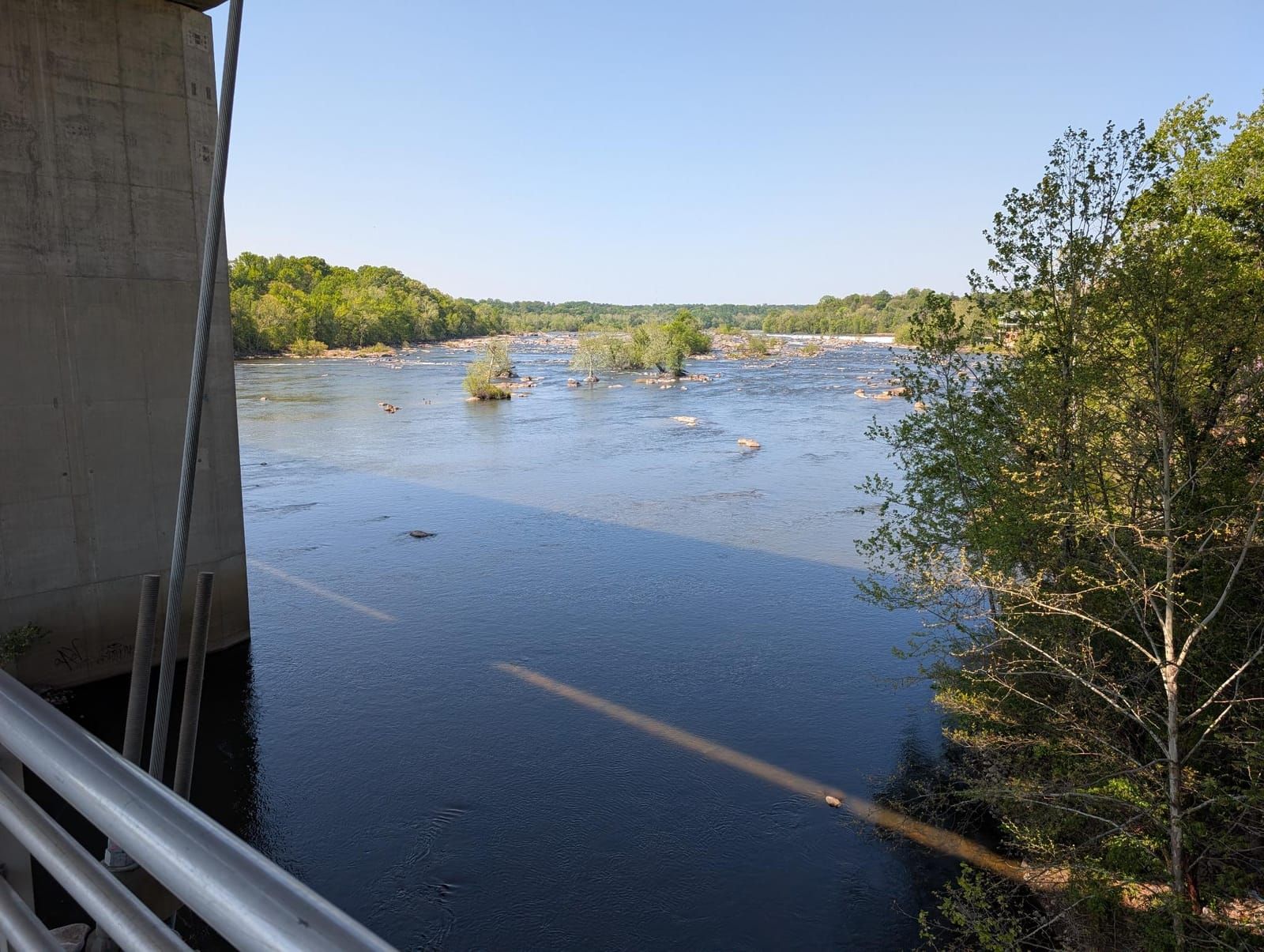 A wide river with rocky outcrops and forested banks stretches across the landscape, viewed from a concrete bridge or structure on the left side under clear blue sky.