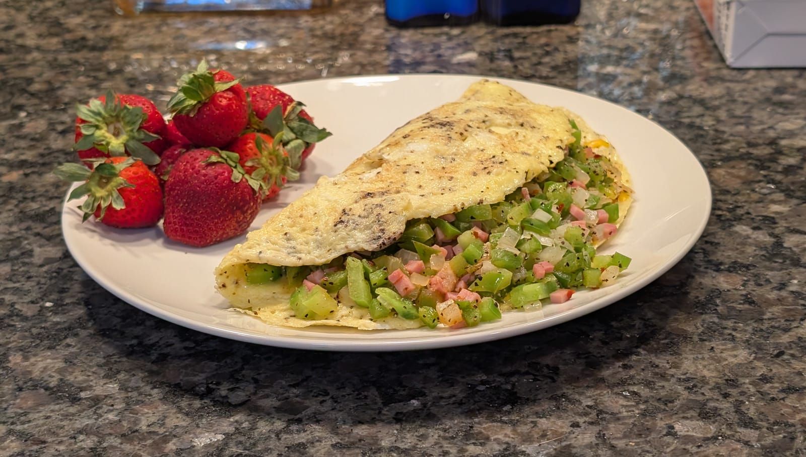 A white plate on a granite countertop holds an omelet filled with diced green peppers and ham, garnished with black pepper, alongside fresh strawberries.