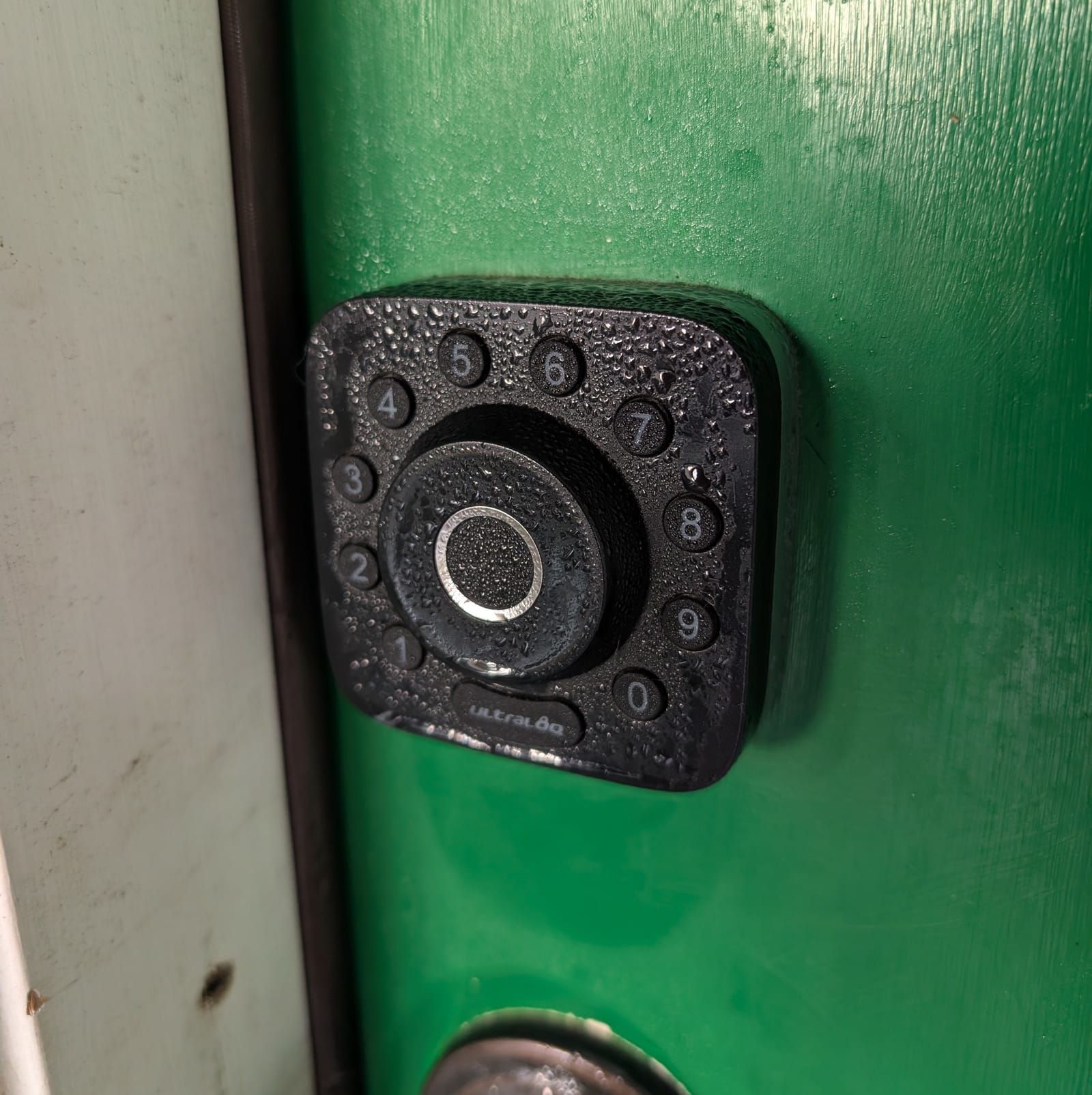 A black rotary telephone dial mounted on a green painted wall, showing numbers 0-9 arranged in a circle around a central metal finger hole, with water droplets covering its surface.