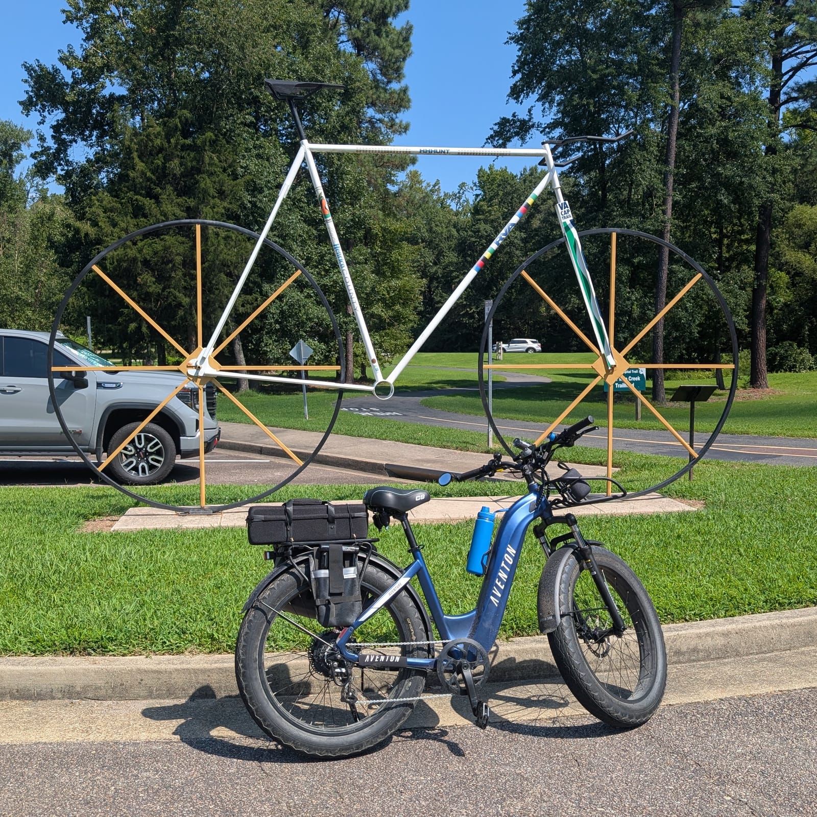 A blue electric bike with a rear cargo rack stands in front of a large sculptural bicycle art installation made of white metal tubing and black wheels with yellow spokes, displayed in a park setting with trees and grass in the background.