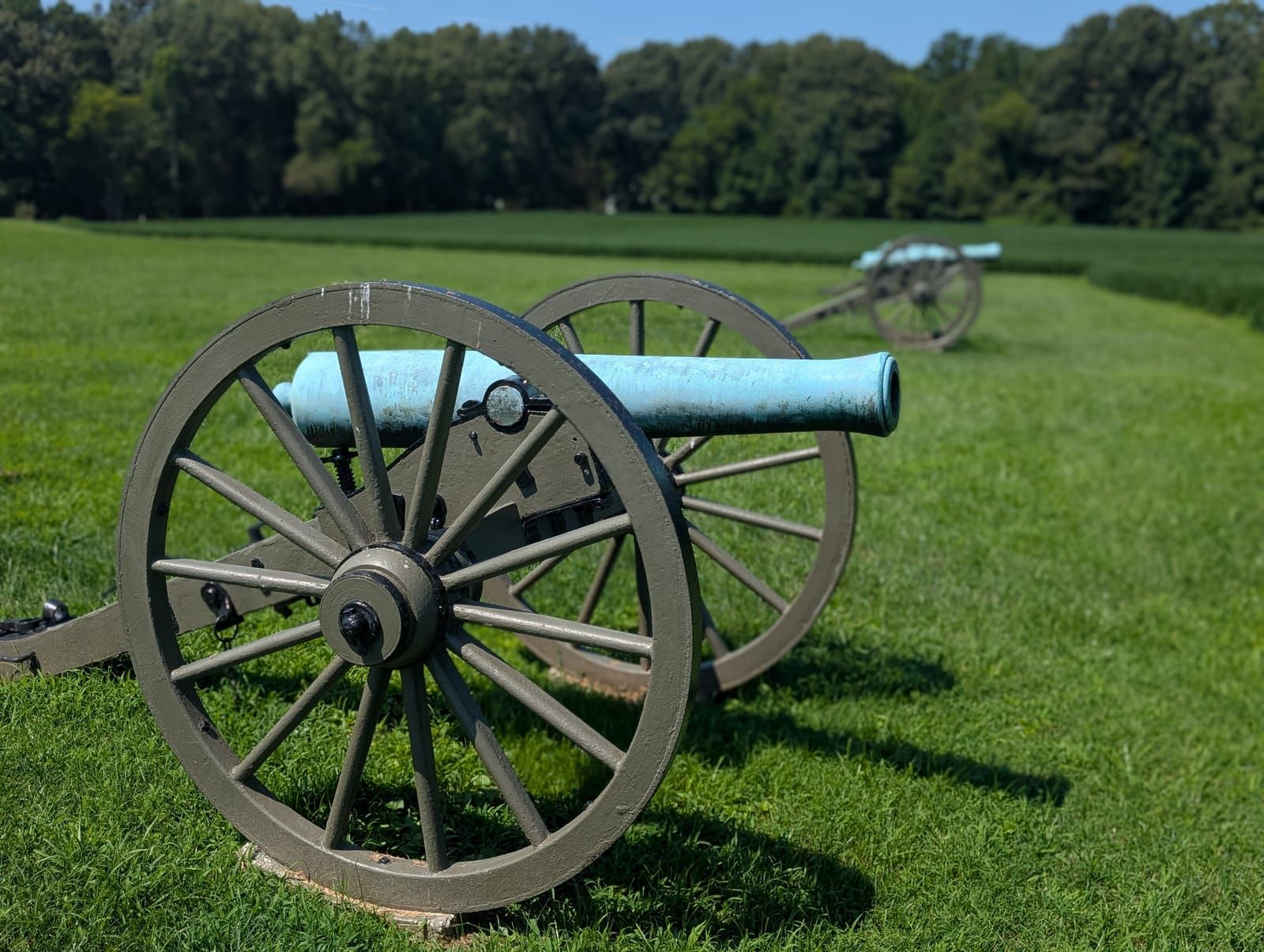 Two Civil War cannons on a mown grass field, the nearer one showing an olive-green wooden carriage with tall spoked wheels and a bronze barrel weathered to pale blue-green. A second cannon sits further back to the right, with a dense treeline and clear blue sky beyond.