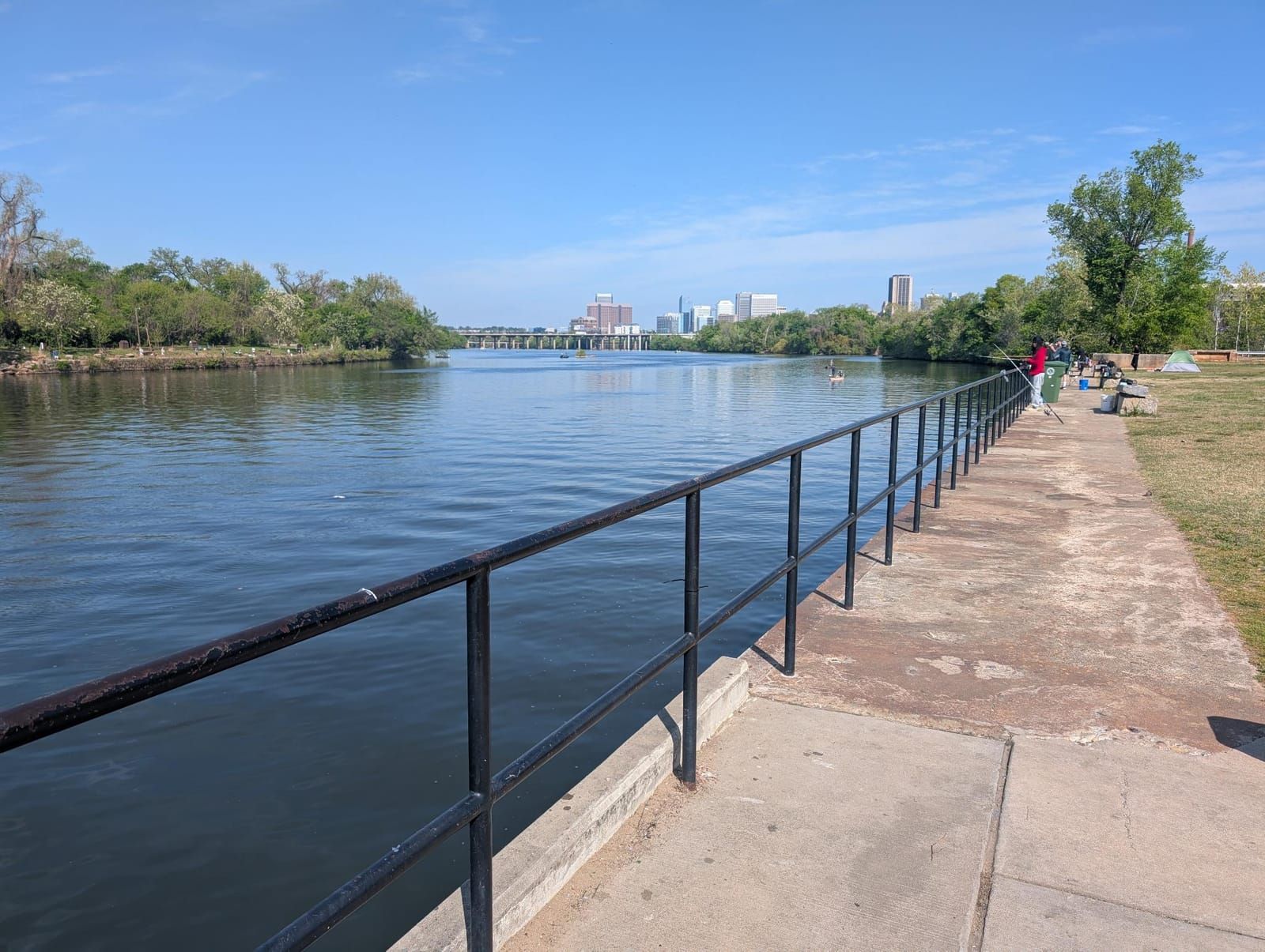 A waterfront promenade with a black metal railing overlooks a calm blue river, with a city skyline visible across the water in the background. Several people are scattered along the grassy park area on the right, enjoying the scenic riverside setting on a clear day.