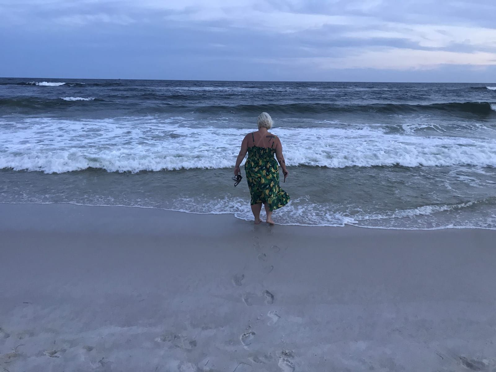 Rachelle walking into the shallow surf wearing a dress and holding her shoes.