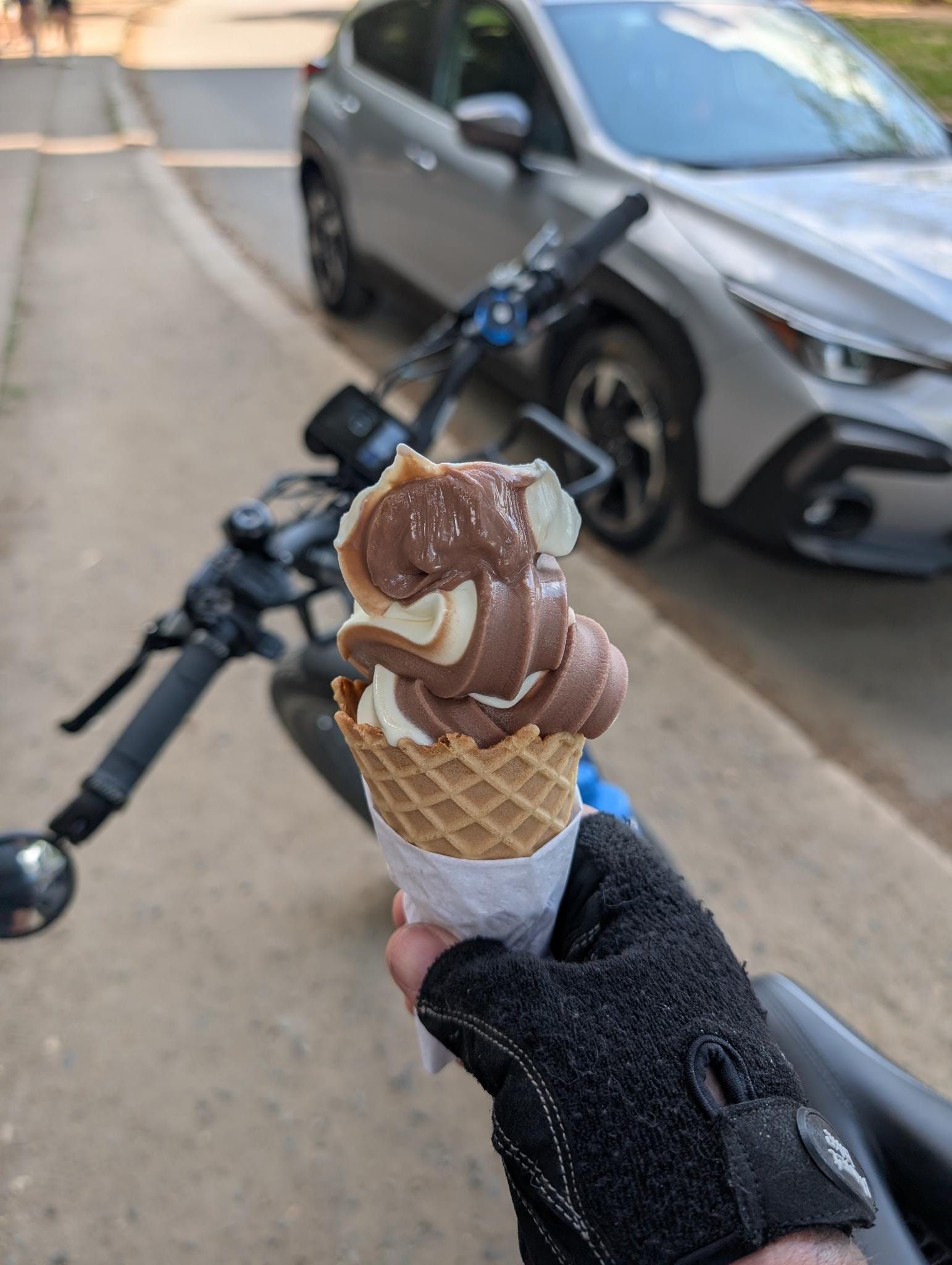 A hand wearing a black glove holds a chocolate and vanilla swirl ice cream cone in a waffle cone. In the blurred background, a bicycle and parked cars are visible on a residential street.