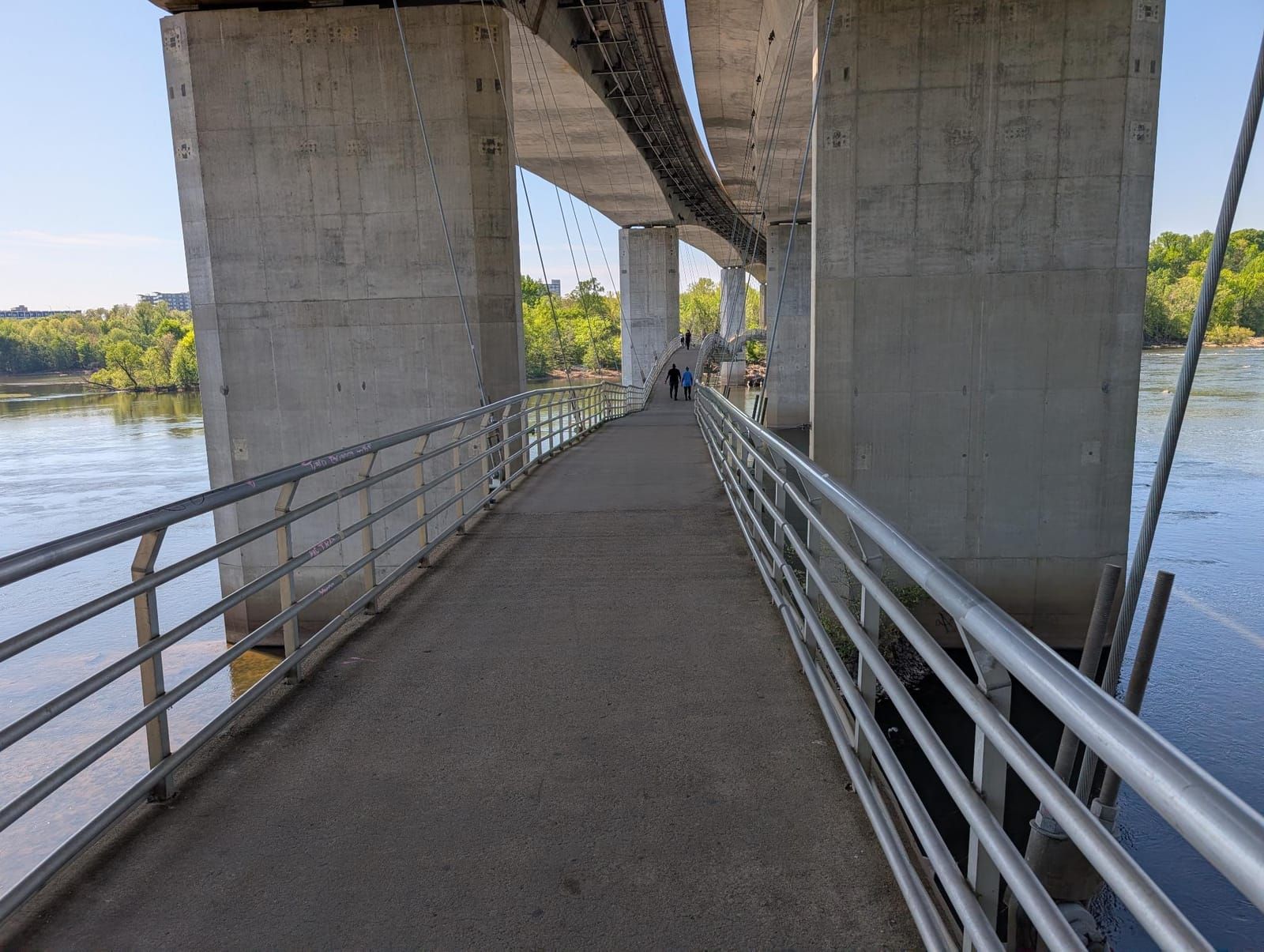 A pedestrian walkway runs between massive concrete bridge pillars spanning a river, with metal railings on both sides and a few people visible in the distance. Green trees line the riverbanks under a clear blue sky.