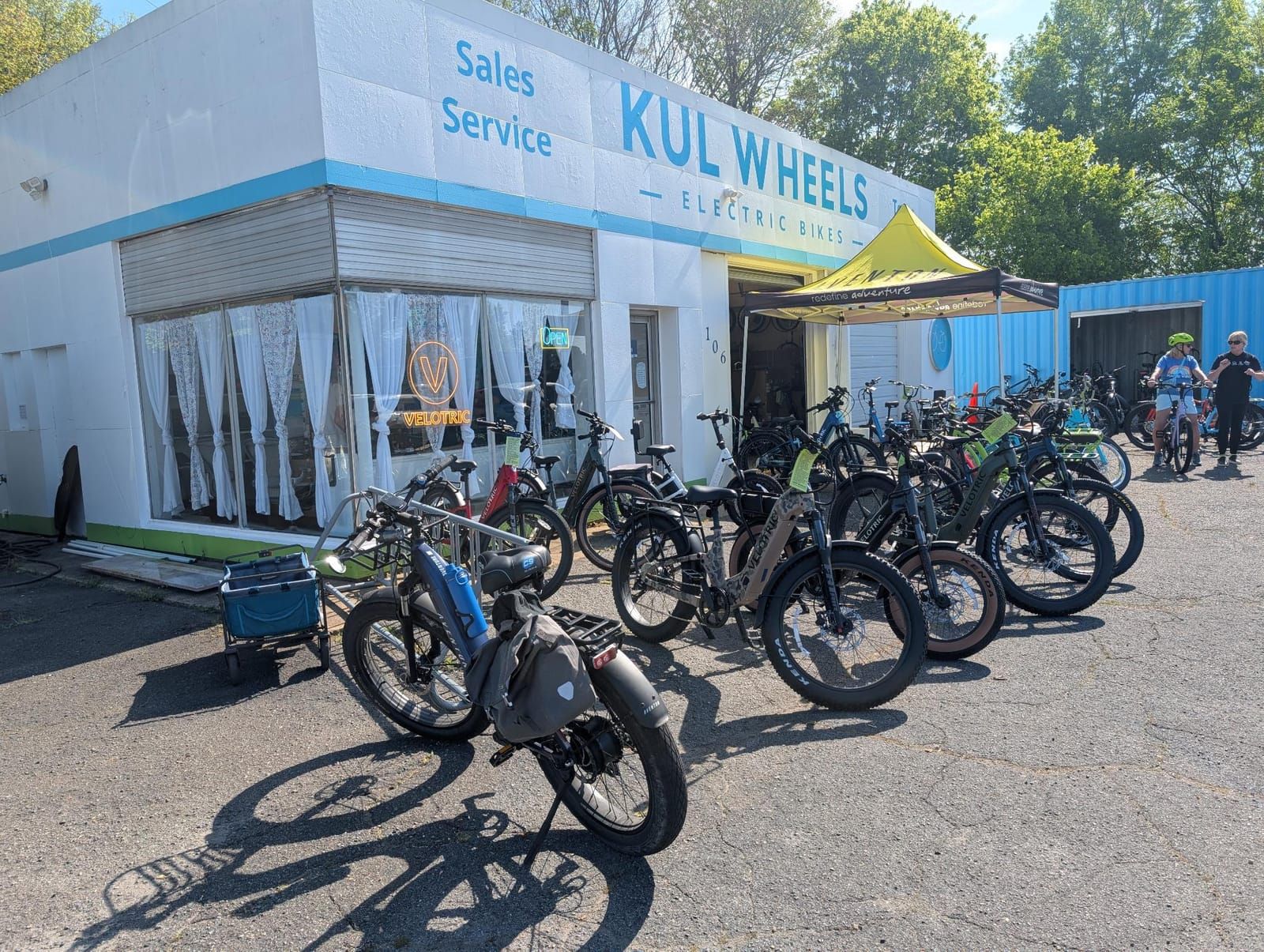 A KUL Wheels electric bikes shop with a white building displaying Sales Service signage. Multiple electric bicycles are parked outside on the pavement, with a yellow tent canopy and customers visible near the entrance.