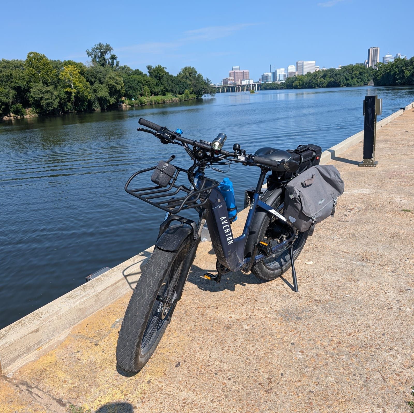 An electric bike with blue and black coloring and a gray cargo bag parked on a concrete pier overlooking a calm river, with a city skyline visible across the water in the background.