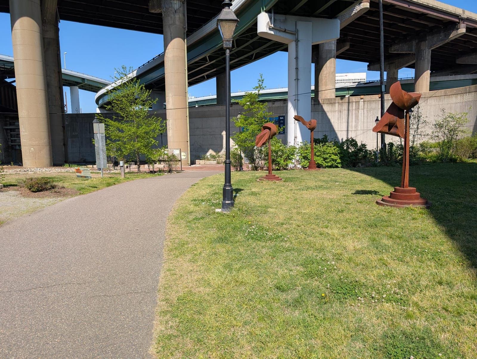 A public park area beneath highway overpasses features a paved pathway, green grass, young trees, and three rust-colored abstract metal sculptures. Concrete pillars support the elevated highways above.