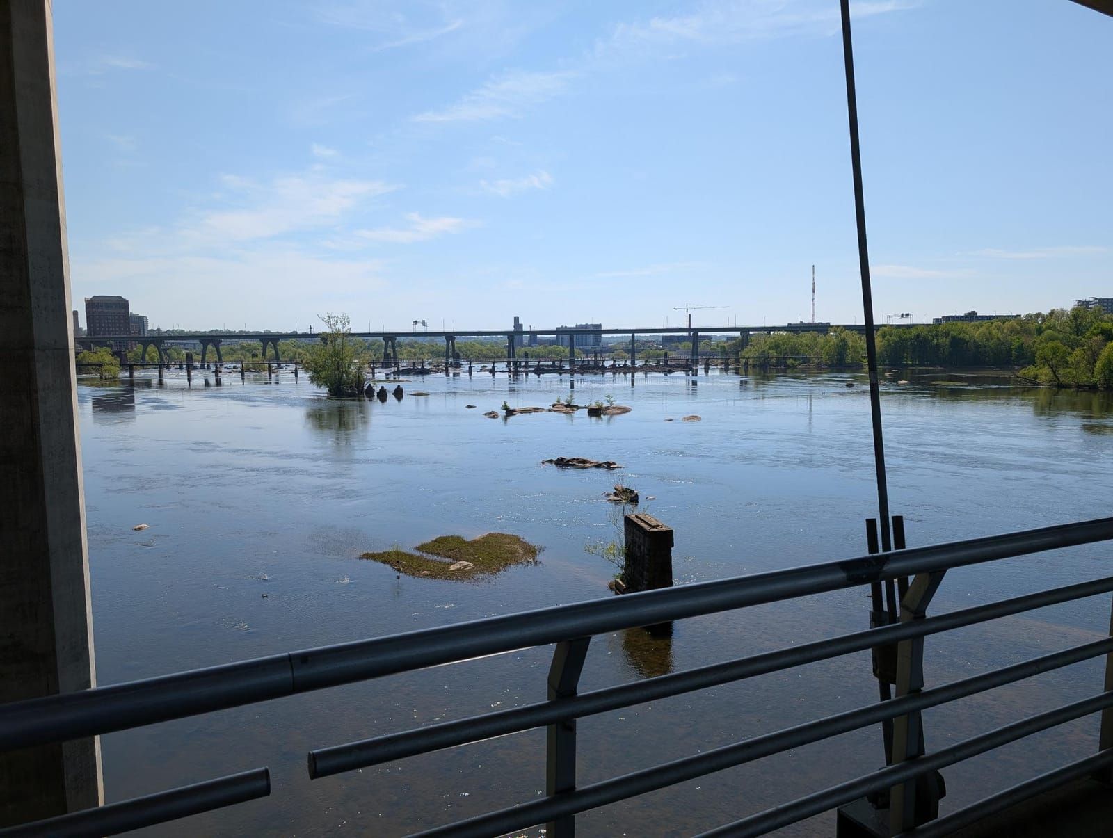 View from a railing overlooking a wide river at flood stage, with exposed tree stumps and vegetation islands visible in the water. A highway bridge spans across the river in the background under a clear blue sky with forested banks on both sides.