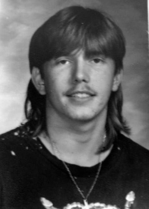 Black and white portrait photograph of a man with long dark hair and a mustache, wearing a dark shirt with a necklace, against a plain gray background.