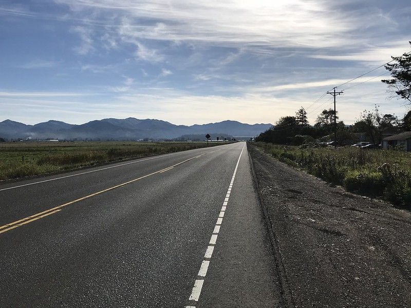 Photo from the side of the road with a long straight road disappearing into the distance with mountains in the background.