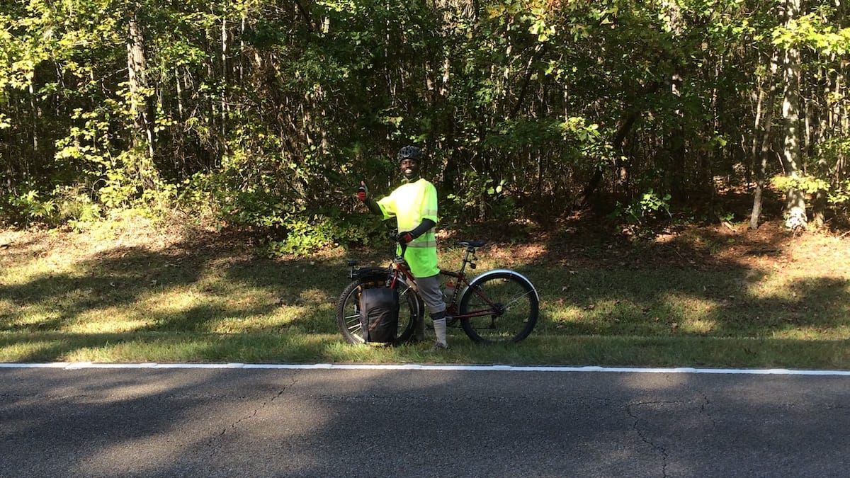 Cyclist wearing bright yellow jersey, standing on the opposite side of the road, waving and smiling.