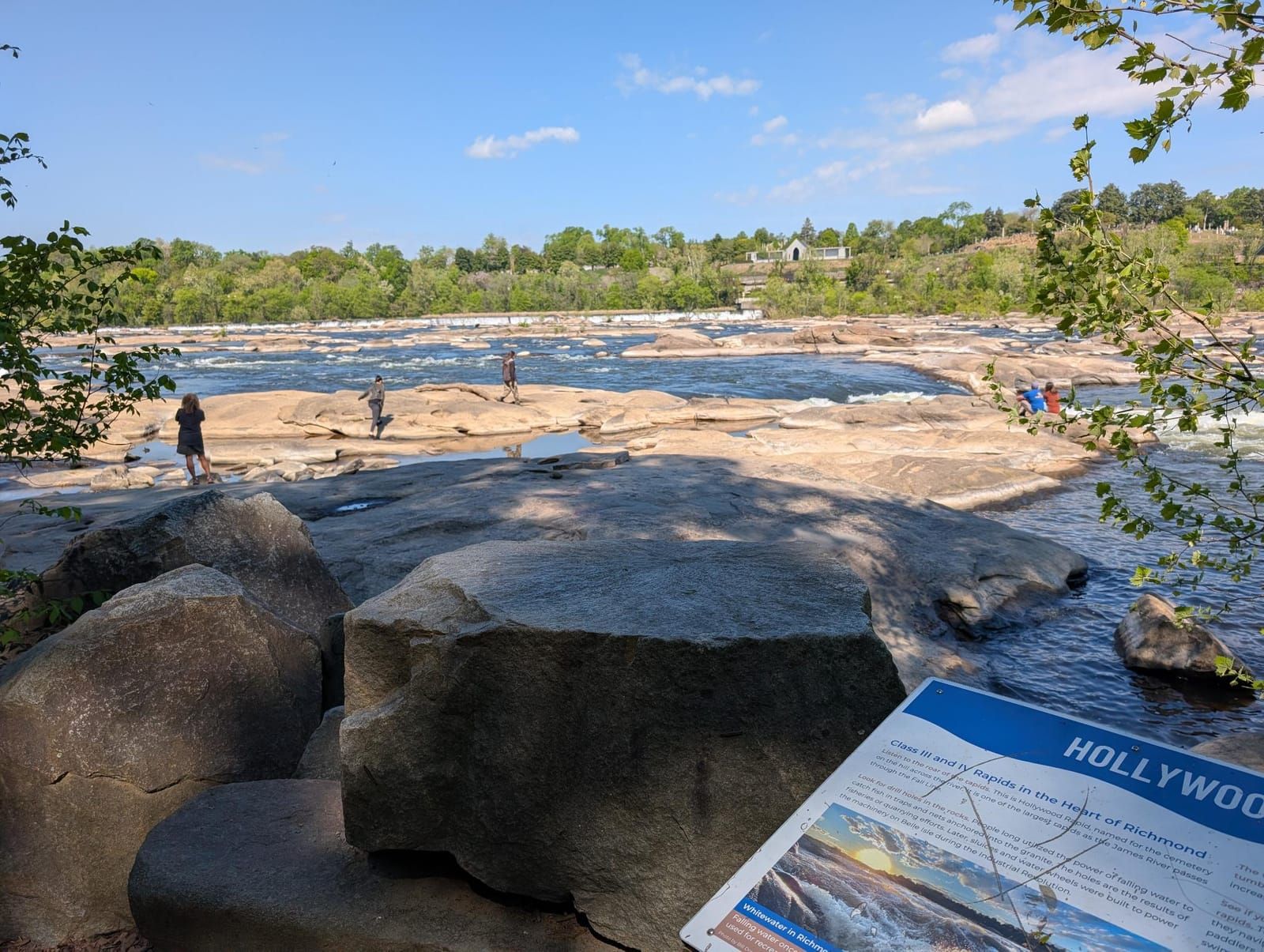 A scenic riverside area with large boulders in the foreground and an informational sign about Hollywood Rapids visible in the lower right. In the background, people explore rocky outcrops along the flowing river, with forested hills and houses visible across the water under a partly cloudy sky.