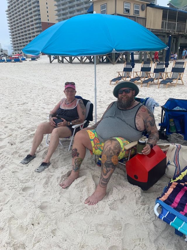 Mom Linda sitting with Mike on the beach under a beach umbrella with coolers and other beach equipment nearby.