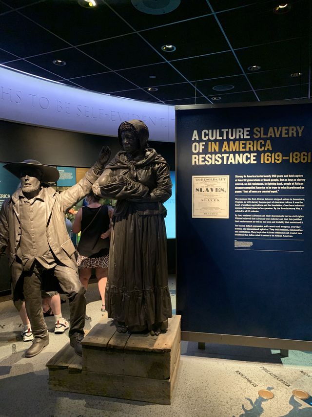 Museum display two statues, one an African woman holding her baby on a literal auction block while a white slaver displays her for sale. Informational sign is to the right.