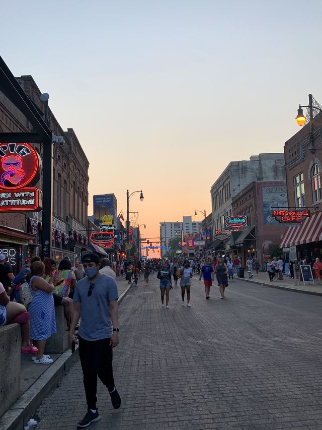 A busy Beale Street at sunset. People are walking in the street and on the sidewalks and bright neon signs advertise the establishments.
