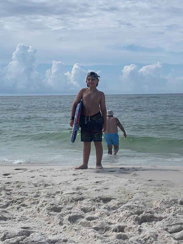 10 year old Carter standing on the beach with a boogie board wearing a hat posing for the camera with the ocean in the background.