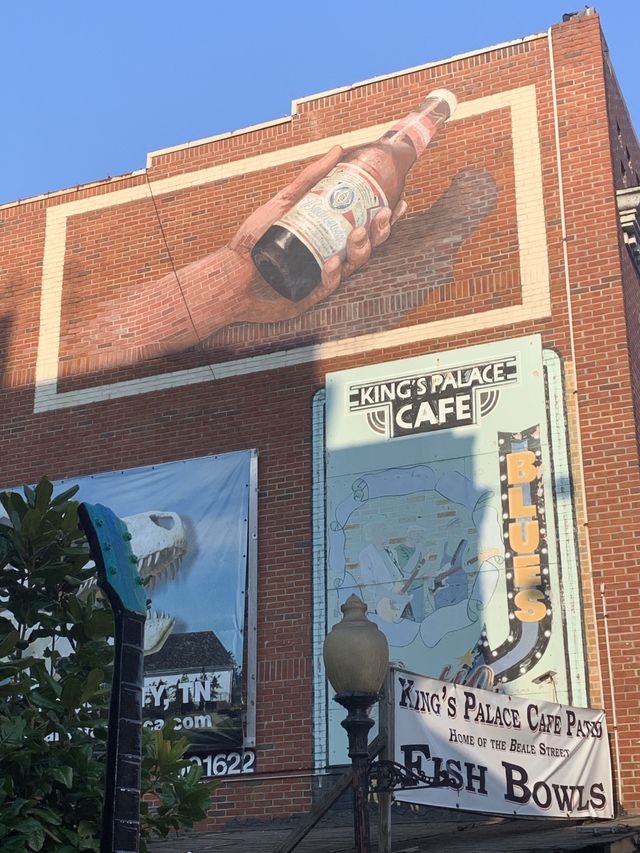 Building mural on the side of Kings Palace Cafe depicting a hand holding a bottle of budweiser with a shadow and 3d effect.