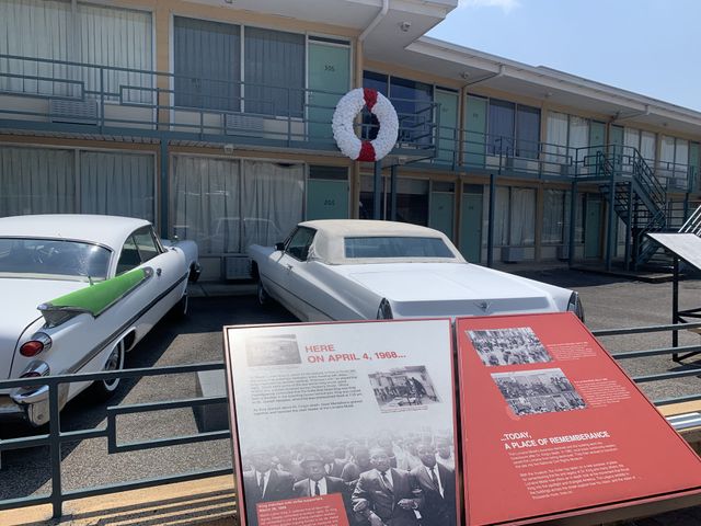 A large white wreath with white stripes is placed on the second level of the Lorraine Motel where Doctor King was killed. In the foreground, 1950s era cars are still parked in front of the preserved motel scene.