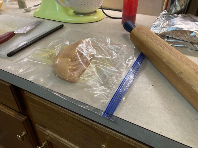 An unprepared chicken breast in a gallon ziplock bag with a large rolling pin laying next to it on the counter.
