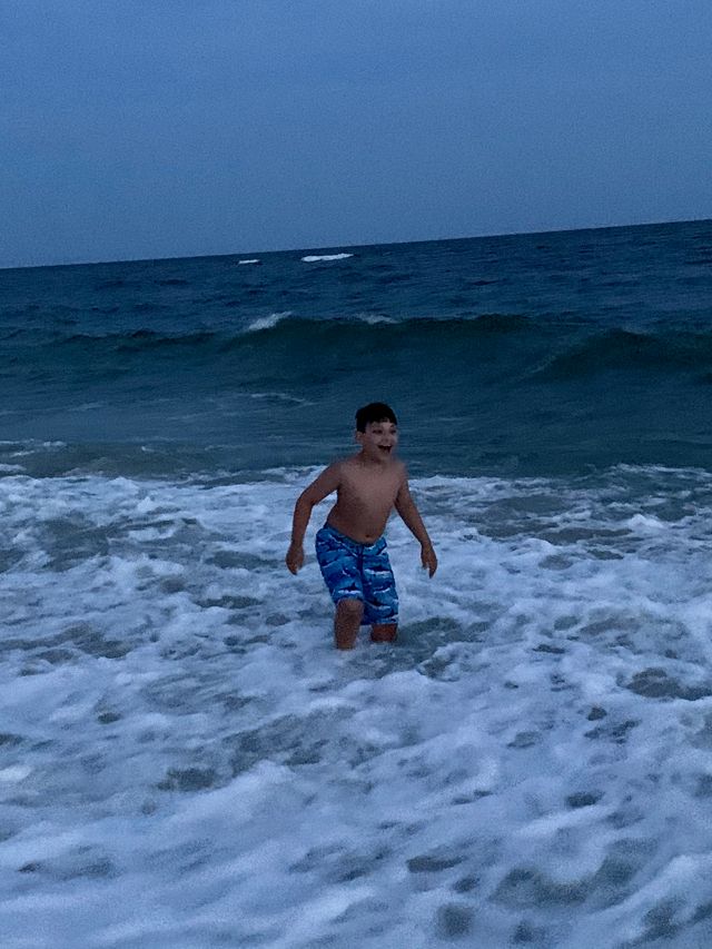 10 year old Carter wearing blue swim tunks entering the evening ocean surf with surprised smile on his face.