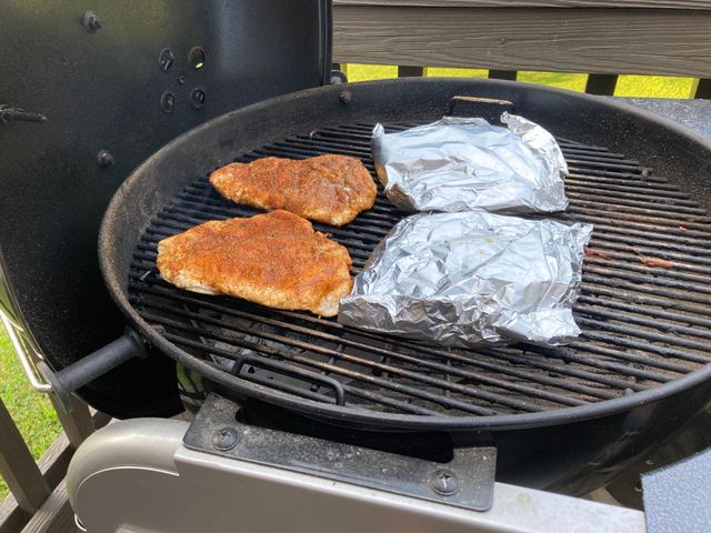 Flattened and rub covered chicken breast on the grill with two foil packets to the side.