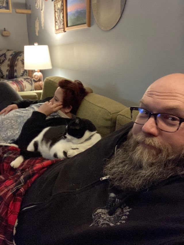 Mike and Rachelle sitting on the couch. Oliver the fat cat lays on Mike.
